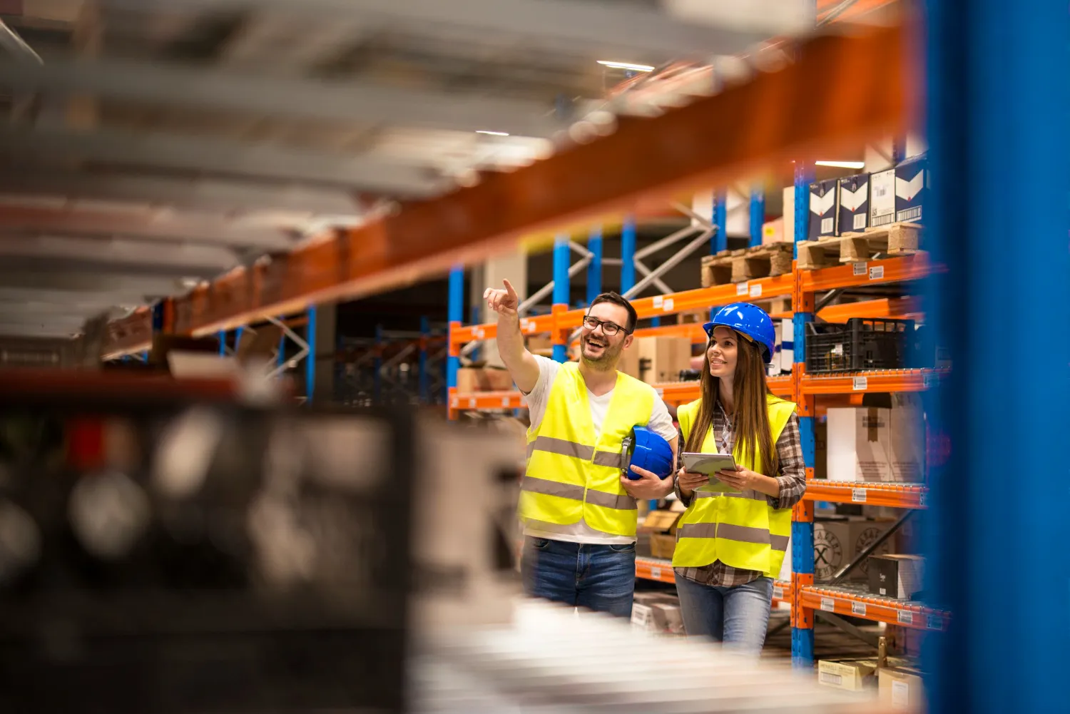 A man and woman wearing high-visibility vests stand in a large warehouse aisle. The man points toward a high shelf while the woman holds a digital tablet and smiles.