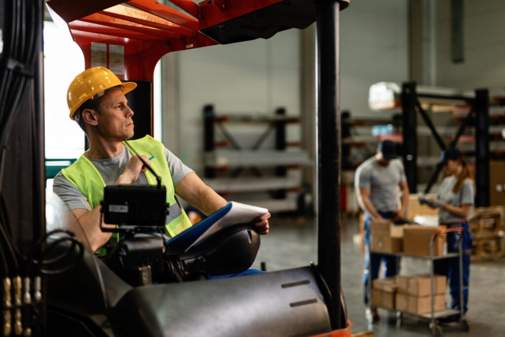 Forklift Safety and Efficiency for a Fortune 500 Automotive Manufacturer 3 Forklift operator in a hi-vis vest operating in a warehouse with pedestrian workers in the background representing forklift pedestrian safety risks in manufacturing environments