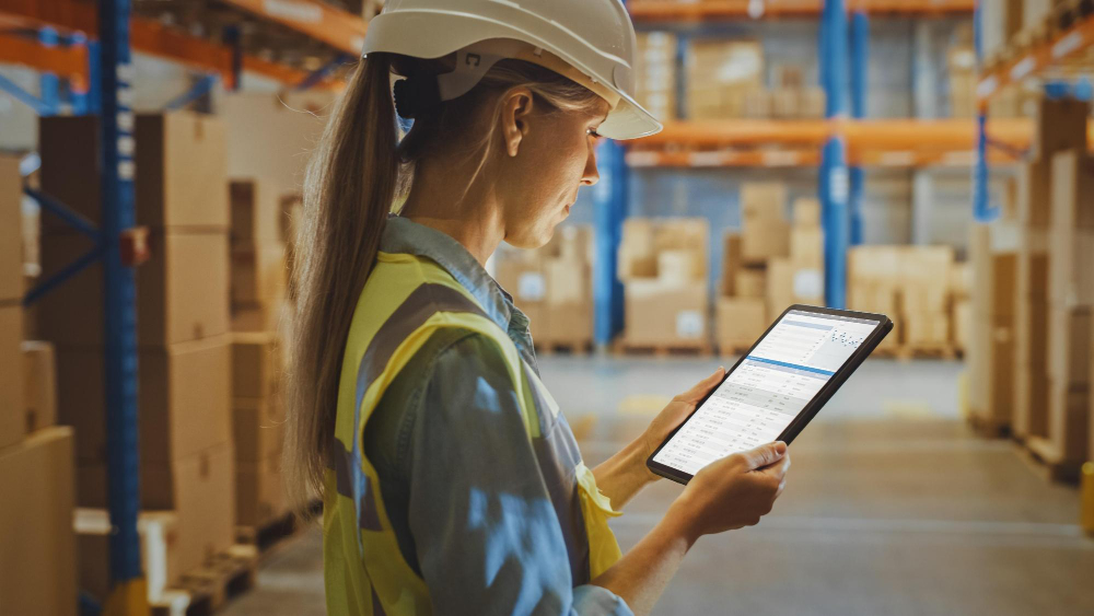 Why Asset Tracking and Connected Worker RTLS Deliver a Multiplier Effect 1 Warehouse manager in a hard hat and hi-vis vest reviewing asset tracking data on a tablet in a large industrial warehouse representing real-time asset management software