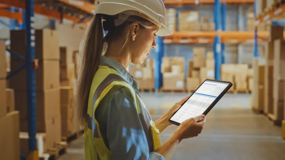 Why Asset Tracking and Connected Worker RTLS Deliver a Multiplier Effect 1 Warehouse manager in a hard hat and hi-vis vest reviewing asset tracking data on a tablet in a large industrial warehouse representing real-time asset management software
